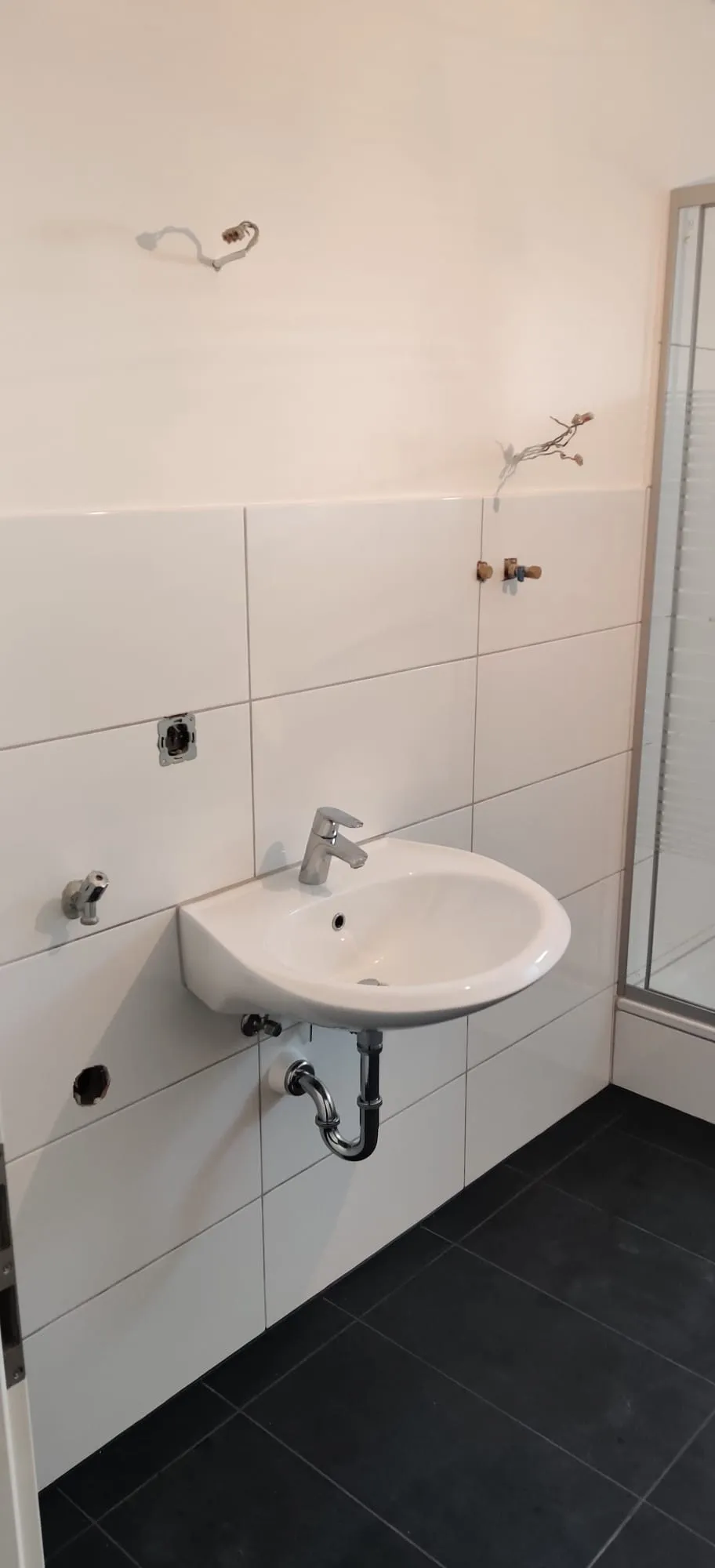 Waschbecken an weiß gefliester Wand mit Waschmaschinenanschlüssen und Duschkabine daneben